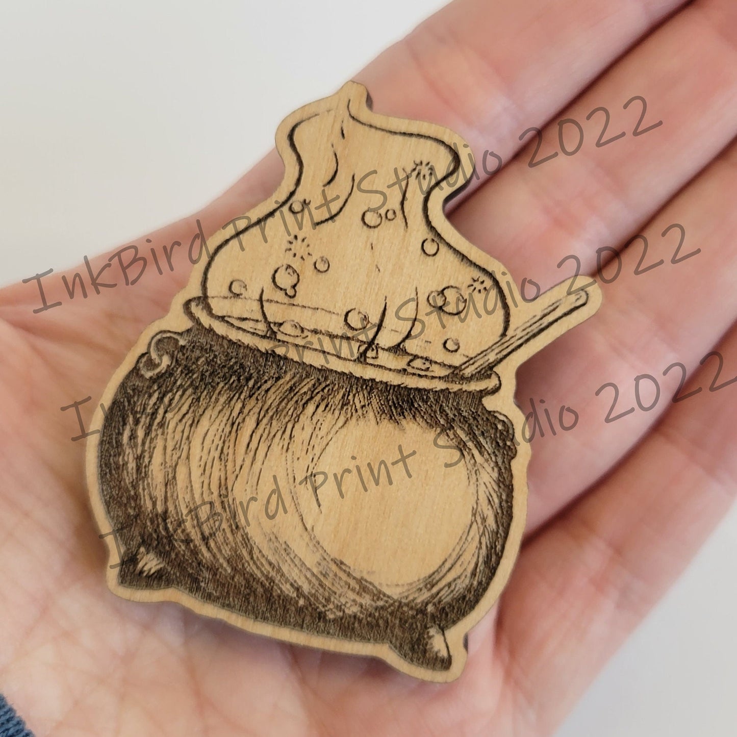 A hand displays InkBird Print Studio’s wood magnet, engraved with intricate bubbling cauldron and spoon art; strong neodymium backing.