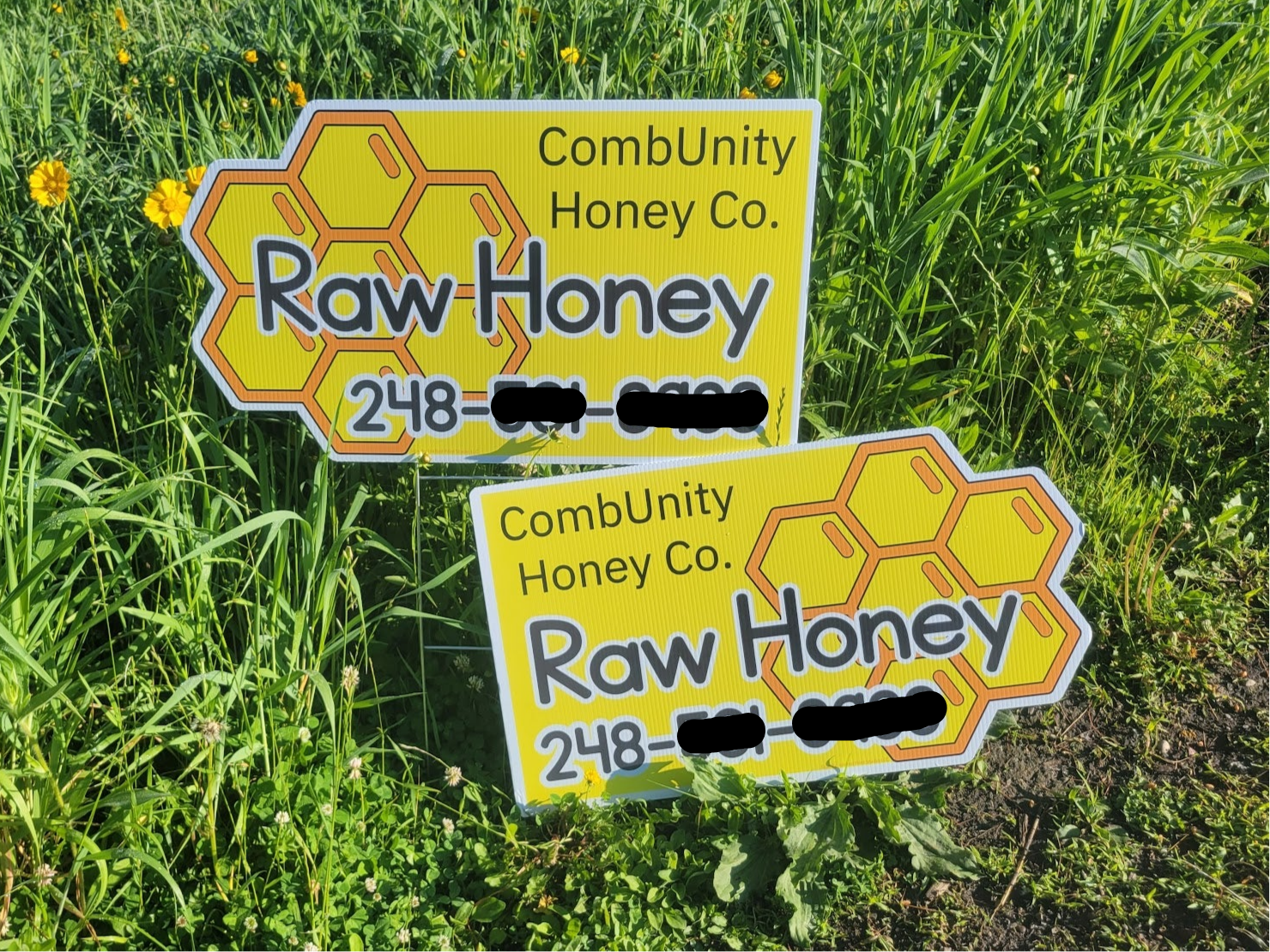 Two custom vinyl yellow and white signs for CombUnity Honey Co. Raw Honey with phone number, staked in green grass with flowers.