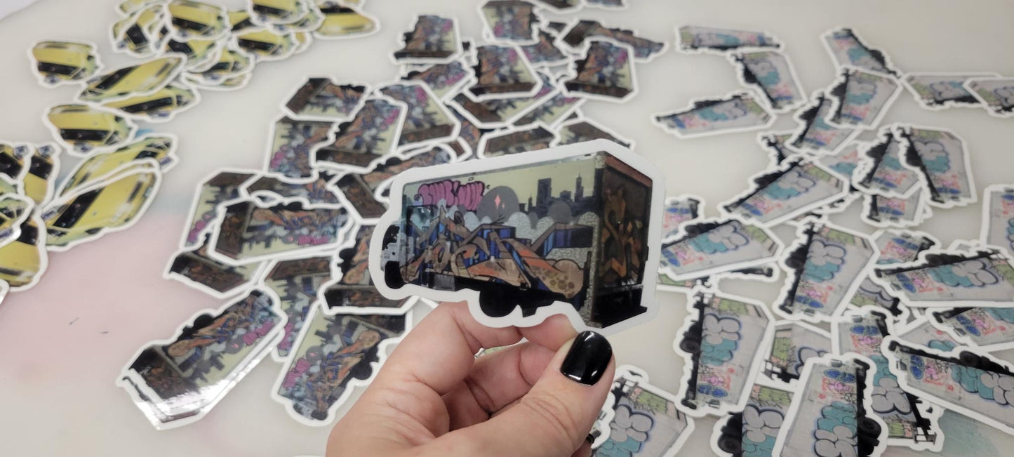 Die cut vinyl sticker of a graffiti truck by InkBird Print Studio, held by hand with black nails; custom stickers on white background.